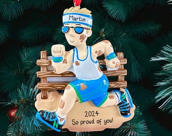 May include: A personalized Christmas ornament featuring a cartoon runner with brown dirt smudges on their face and body. The runner is wearing a white tank top, blue shorts, a blue headband with the name "Martin" written on it, and sunglasses. The runner is running over a brown wooden fence. The ornament also includes the text "2024" and "So proud of you!"