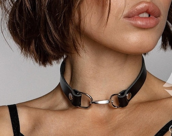 May include: A black leather choker featuring a central silver-toned metal ring and hardware, worn around the neck. The choker is paired with a black strappy harness garment. A white graphic of lips with the text BdsmCherry is visible.