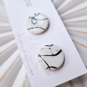 Könnte beinhalten: Ein Paar weiße, runde Ohrringe mit einem schwarzen, abstrakten Design. Die Ohrringe werden auf einer weißen Karte mit dem Text "KELANI HANDMADE JEWELLERY" präsentiert. Der Hintergrund ist eine weiche, neutrale Farbe.