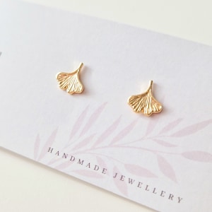 Könnte beinhalten: Ein Paar goldfarbene Ohrstecker in Form von Ginkgoblättern. Die Ohrringe werden auf einer weißen Karte mit dem Text "HANDMADE JEWELLERY" präsentiert. Die Ohrringe haben eine detaillierte, strukturierte Oberfläche.