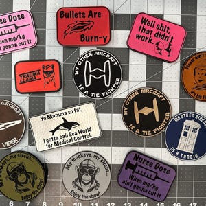 May include: A collection of 11 embroidered patches with various designs and sayings. The patches are on a gray grid background. Some of the patches include: "Nurse Dose When mg/kg ain't gonna cut it", "Bullets Are Burn-y", "My Other Aircraft Is A Tie Fighter", "My Other Aircraft Is A Viper", "Yo Mamma So Fat, I Gotta Call Sea World For Medical Control", "My Monkeys, My Circus... Enjoy The Show!" and "Please Don't Talk To The Pilot, It's His First Day".