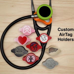 May include: A black stethoscope with a green and orange chest piece, surrounded by several custom AirTag holders featuring anatomical designs like a heart, lungs, brain, liver, and kidneys, with text reading Custom AirTag Holders.