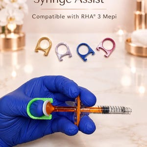 Può includere: ThumbMate Syringe Assist compatibile con RHA 3 Mepi. Una mano con guanto blu tiene una siringa con un anello per pollice verde brillante. Quattro anelli aggiuntivi in oro, argento, blu e magenta sono mostrati sopra la siringa.