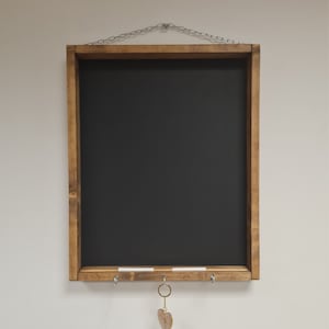 May include: A wooden framed chalkboard with a black surface, three hooks, and a small heart-shaped keychain hanging from one of the hooks. The chalkboard is hanging from a chain.