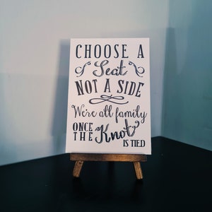 May include: White sign with black text that reads "Choose a Seat Not a Side We're all family once the Knot is tied". The sign is on a small wooden easel.