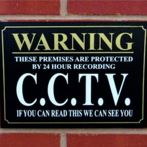 May include: Black and gold sign with the text "WARNING THESE PREMISES ARE PROTECTED BY 24 HOUR RECORDING C.C.T.V. IF YOU CAN READ THIS WE CAN SEE YOU"