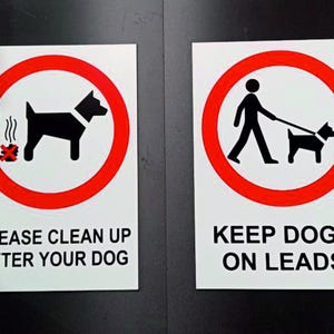 May include: Two white rectangular signs with black text and red circular symbols. One sign reads "PLEASE CLEAN UP AFTER YOUR DOG" and depicts a dog and waste symbol. The other reads "KEEP DOGS ON LEADS" and shows a person walking a dog.