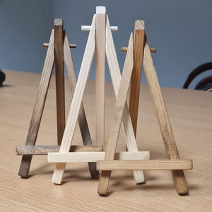 May include: Three wooden easels, one natural wood and two stained brown, are arranged together on a light wood surface. The easels are small and designed to hold miniature paintings or photos.