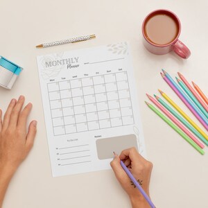Op de afbeelding: Een witte maandelijkse planner met een bloemenontwerp, een to-dolijst en een notitiegedeelte. Een hand schrijft op de planner met een paarse pen. Er liggen kleurpotloden en een kop koffie op de tafel.