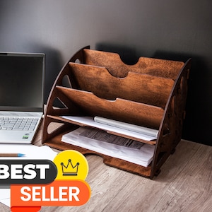 Peut inclure: Un organiseur de bureau en bois avec quatre compartiments inclinés, teinté d'un brun chaud. Il contient des papiers et un stylo. Un ordinateur portable et les mots "BEST SELLER" sont également visibles.