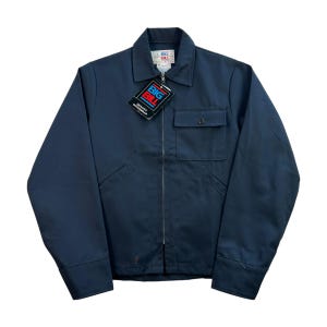 Op de afbeelding: Een marineblauwe werkjas met een ritssluiting over de hele lengte, een borstzak en twee zijzakken. De jas heeft een kraag en lange mouwen met manchetten met knopen. Een label met het "BIG BILL"-logo is bevestigd.