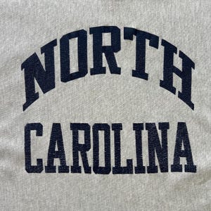 Puede incluir: Primer plano de una tela gris claro con las palabras "NORTH CAROLINA" impresas en azul marino. La palabra "NORTH" est&aacute; arqueada sobre "CAROLINA". El texto tiene un aspecto vintage y desgastado.