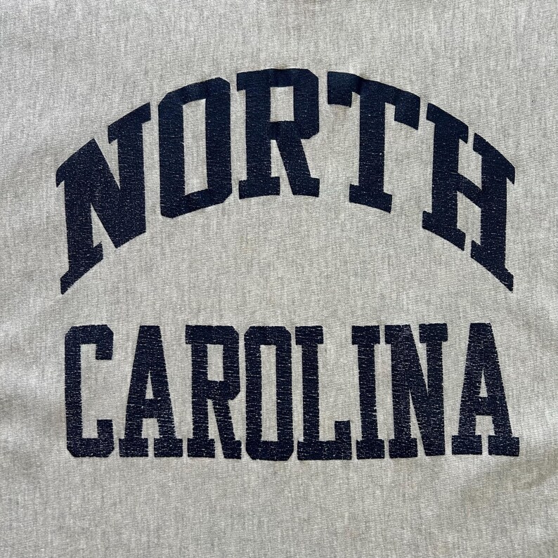 Puede incluir: Primer plano de una tela gris claro con las palabras "NORTH CAROLINA" impresas en azul marino. La palabra "NORTH" est&aacute; arqueada sobre "CAROLINA". El texto tiene un aspecto vintage y desgastado.