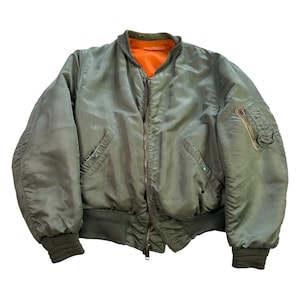May include: An olive green bomber jacket with an orange lining. Features a zip closure, two front pockets, and a zipped pocket on the left sleeve. The cuffs and waistband are ribbed.