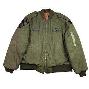 May include: An olive green bomber jacket with a ribbed collar, cuffs, and hem. The jacket has the name "MILLER" and "U.S. ARMY" printed on the front. Patches are visible on the sleeves, and there is a zipped pocket. The lining is orange.