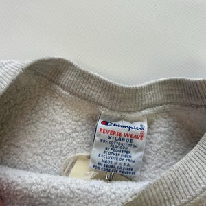 Puede incluir: Primer plano de una sudadera Champion Reverse Weave gris. La etiqueta dice "X-LARGE" y enumera la composici&oacute;n de la tela: 89% algod&oacute;n, 8% poli&eacute;ster y 3% otras fibras. La etiqueta tambi&eacute;n dice "Made in U.S.A."