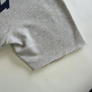 Puede incluir: Primer plano de una sudadera gris de manga corta. El borde sin rematar de la manga es visible. La tela es de color gris claro.