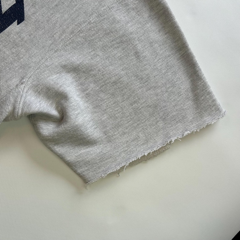 Puede incluir: Primer plano de una sudadera gris de manga corta. El borde sin rematar de la manga es visible. La tela es de color gris claro.
