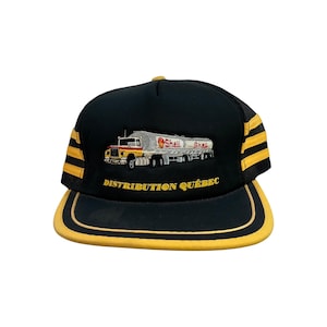 May include: Black trucker hat with yellow accents and a curved brim. The hat features an embroidered image of a Shell tanker truck and the text "DISTRIBUTION QUÉBEC". The hat has a mesh back and a snap closure.