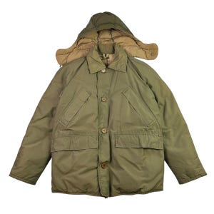 Puede incluir: Abrigo de invierno verde oliva con capucha desmontable. El abrigo tiene una botonadura frontal, dos bolsillos en el pecho y dos bolsillos inferiores. La capucha está forrada con una tela beige más clara. Diseñado para el frío.