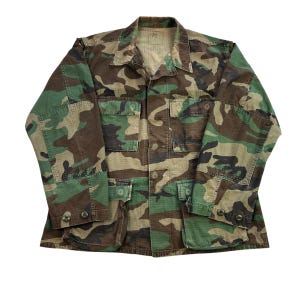 Könnte beinhalten: Eine Tarnmusterjacke in Grün-, Braun- und Beigetönen. Die Jacke hat eine Knopfleiste, vier Vordertaschen und geknöpfte Manschetten. Der Kragen ist im klassischen Stil gehalten. Die Jacke hat die Größe Medium.
