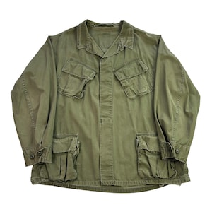 May include: An olive green military-style jacket with four front pockets and two buttoned cuffs. The jacket has a collared neckline and a button-up front. The fabric appears to be a durable, woven material.