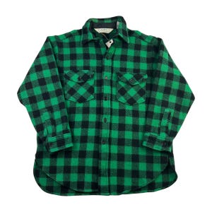 May include: Green and black checked button-down shirt with two chest pockets. The shirt features a classic collar and buttoned cuffs. The fabric has a thick, woven texture. The shirt is a classic lumberjack style.