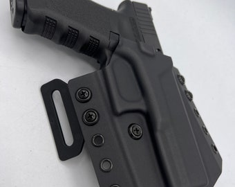 Puede incluir: Una funda de polímero negro que contiene una pistola semiautomática negra. La funda cuenta con un accesorio para cinturón en el lateral, asegurado con varios tornillos negros y ojales metálicos a lo largo del borde, ofreciendo un ajuste moldeado.