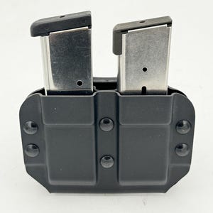 Może przedstawiać: Czarny podwójny uchwyt na magazynki z dwoma srebrnymi magazynkami. Magazynki mają czarne wierzchy i są bezpiecznie trzymane w uchwycie. Uchwyt jest wykonany z wytrzymałego materiału i posiada kilka nitów.