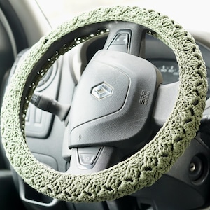 以下が含まれることがあります： セージグリーンの、かぎ針編みのステアリングホイールカバー。カバーは黒いステアリングホイールに巻き付けられており、かぎ針編みの模様が見えます。ステアリングホイールは車両のダッシュボードの一部です。