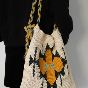 May include: A white crocheted tote bag with a geometric design in black, green, and yellow. The bag has a braided strap in green and yellow.
