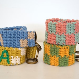 Może przedstawiać: Trzy breloki wykonane na szydełku z różnymi wzorami i kolorami. Pierwszy brelok jest niebieski i beżowy z wyhaftowanym zielonym napisem "KAY". Drugi brelok jest różowy i zielony w kratkę. Trzeci brelok jest żółty i zielony w kratkę.