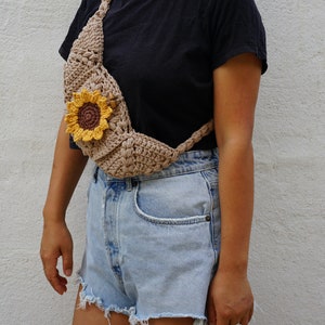 Puede incluir: Una riñonera de crochet con forma de girasol y una correa marrón. La riñonera se lleva sobre el hombro y alrededor de la cintura.