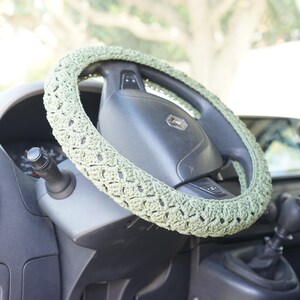 Op de afbeelding: Een lichtgroene, gehaakte stuurwielhoes is te zien op een zwart stuurwiel. De hoes heeft een gestructureerd, open weefpatroon. Het dashboard en andere interieuronderdelen van de auto zijn zichtbaar op de achtergrond.