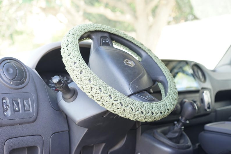 Op de afbeelding: Een lichtgroene, gehaakte stuurwielhoes is te zien op een zwart stuurwiel. De hoes heeft een gestructureerd, open weefpatroon. Het dashboard en andere interieuronderdelen van de auto zijn zichtbaar op de achtergrond.
