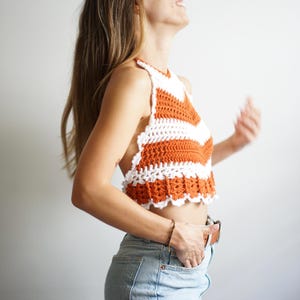 May include: A white and orange crochet halter top with a scalloped hem. The top is made with a simple stitch pattern and has a relaxed fit.