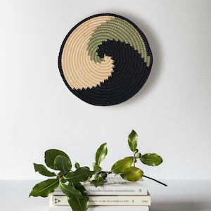 May include: A black, green, and beige crocheted wall hanging in the shape of a spiral. The wall hanging is hanging on a white wall. There are three books stacked below the wall hanging. The top book has the title "Nouvelles histoires extraordinaires" on the spine. The middle book has the title "MAZETTI LE MEC DE LA TOMBE D'À CÔTÉ" on the spine. The bottom book has the title "Ovide Les Métamorphoses" on the spine.