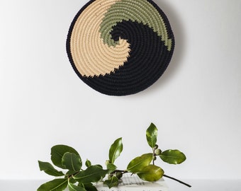 Op de afbeelding: Een zwarte, groene en beige gehäkelde wanddecoratie in de vorm van een spiraal. De wanddecoratie hangt aan een witte muur. Er liggen drie boeken op elkaar onder de wanddecoratie. Het bovenste boek heeft de titel "Nouvelles histoires extraordinaires" op de rug. Het middelste boek heeft de titel "MAZETTI LE MEC DE LA TOMBE D'À CÔTÉ" op de rug. Het onderste boek heeft de titel "Ovide Les Métamorphoses" op de rug.