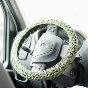 Op de afbeelding: Een gehaakte stuurwielhoes in een lichtgroene tint. De hoes is gewikkeld rond een zwart stuurwiel, met het dashboard en het interieur van het voertuig zichtbaar op de achtergrond. Het haakpatroon voegt textuur en een handgemaakte esthetiek toe aan het interieur van de auto.