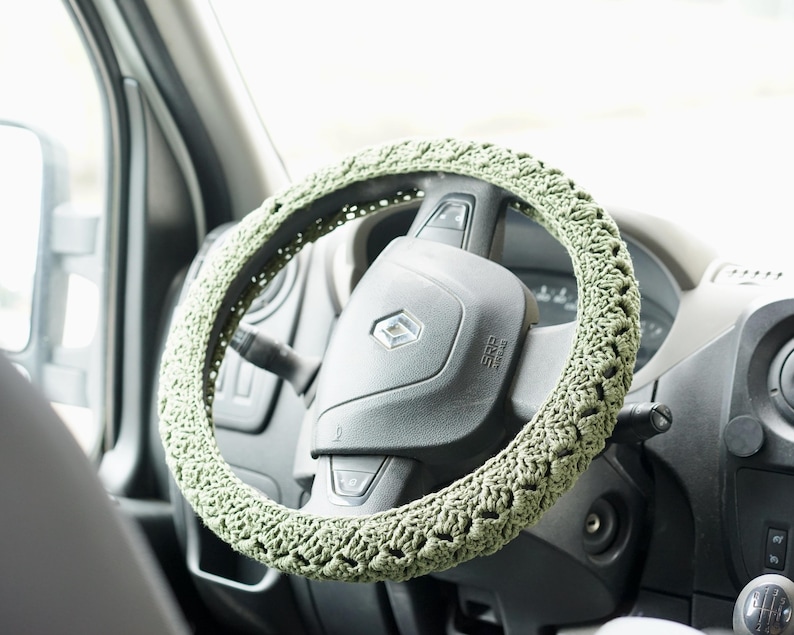 Op de afbeelding: Een gehaakte stuurwielhoes in een lichtgroene tint. De hoes is gewikkeld rond een zwart stuurwiel, met het dashboard en het interieur van het voertuig zichtbaar op de achtergrond. Het haakpatroon voegt textuur en een handgemaakte esthetiek toe aan het interieur van de auto.