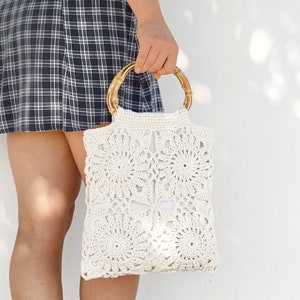 Puede incluir: Un bolso de mano de ganchillo color crema con un asa de bambú. El bolso presenta un patrón floral y una forma rectangular. El asa es de color marrón claro. El bolso lo sostiene una persona que lleva una falda a cuadros.