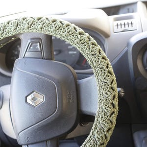 Op de afbeelding: Een saliegroene gehaakte stuurwielhoes. De hoes is gewikkeld rond een zwart stuurwiel met een zilveren embleem. Het interieur van het voertuig is zichtbaar op de achtergrond.