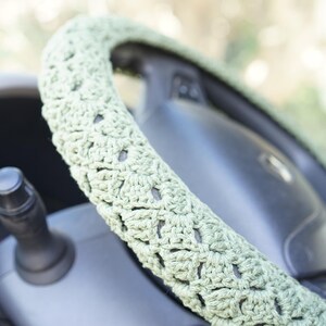 Op de afbeelding: Close-up van een lichtgroene gehaakte stuurwielhoes. De hoes is gewikkeld rond een zwart stuurwiel, met een deel van het dashboard en de richtingaanwijzerhendel zichtbaar. De gehaakte textuur is zichtbaar.