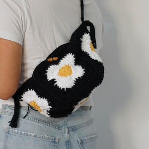 May include: A black crocheted bag with white and yellow daisy-shaped designs. The bag has a long strap and a tassel on the bottom.