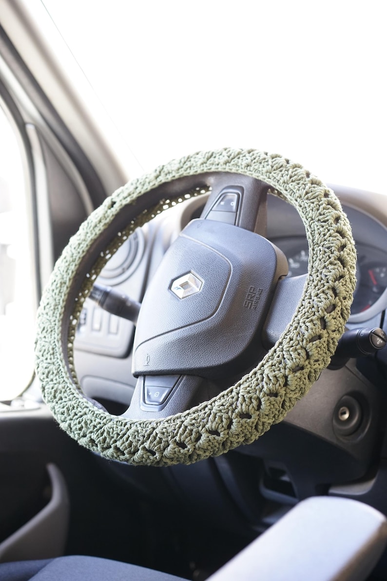 Op de afbeelding: Een saliegroene, gehaakte stuurwielhoes. De hoes is gewikkeld rond een zwart stuurwiel, met het Renault-logo zichtbaar. Het haakpatroon is zichtbaar en voegt textuur toe aan het wiel.