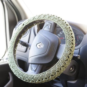 Op de afbeelding: Een gehaakte stuurwielhoes in licht olijfgroen. De hoes zit op een zwart stuurwiel in een voertuig. Het Renault-logo is zichtbaar in het midden van het stuurwiel.