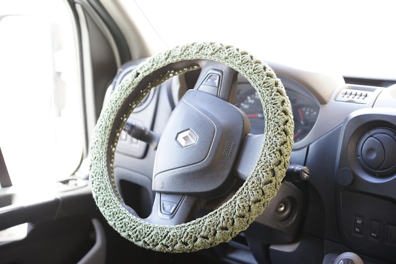 Op de afbeelding: Een gehaakte stuurwielhoes in licht olijfgroen. De hoes zit op een zwart stuurwiel in een voertuig. Het Renault-logo is zichtbaar in het midden van het stuurwiel.