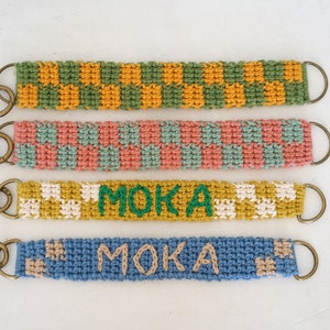 May include: Four crocheted keychains with gold metal rings. The keychains are in different colours and patterns, including green and yellow checked, pink and green checked, white and yellow checked, and blue with beige accents. Two of the keychains have the word "MOKA" embroidered in green.