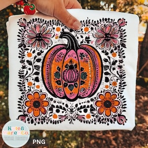 May include: A white t-shirt featuring a central orange and pink pumpkin graphic surrounded by a black floral and vine border. The design includes four orange flowers in the corners and small orange dots, with a circular logo in the bottom left corner.