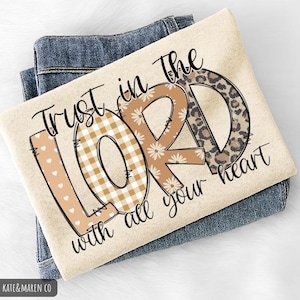 May include: A beige t-shirt with the words "Trust in the LORD with all your heart" in black script. The letters "L" and "O" are decorated with a heart and gingham pattern, and the letter "D" has a leopard print design. The shirt is folded on top of blue jeans.